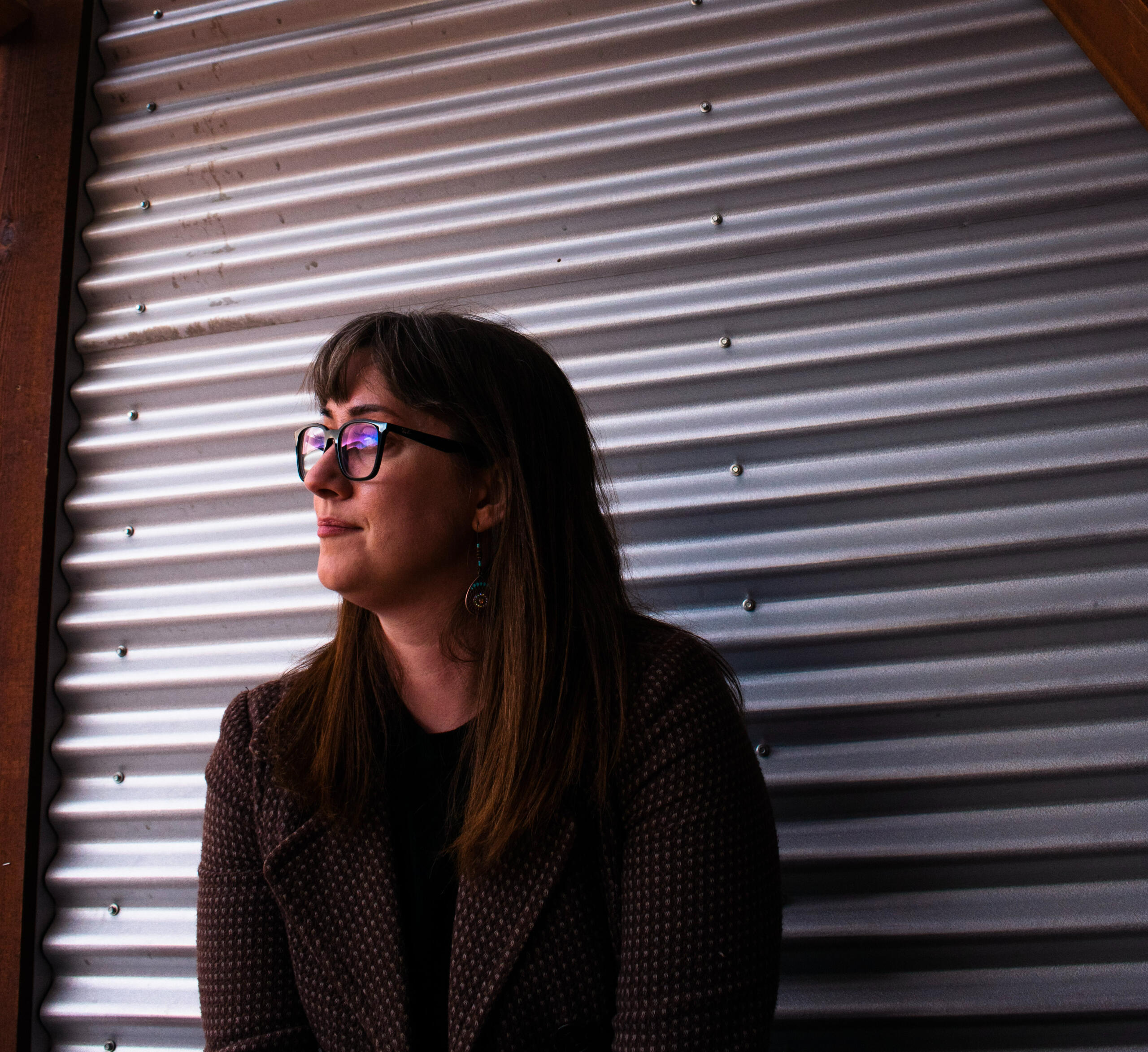 Jess Wesley author A photo of a woman with long brown hair and glasses looking away from the camera against a backdrop of corrugated metal.